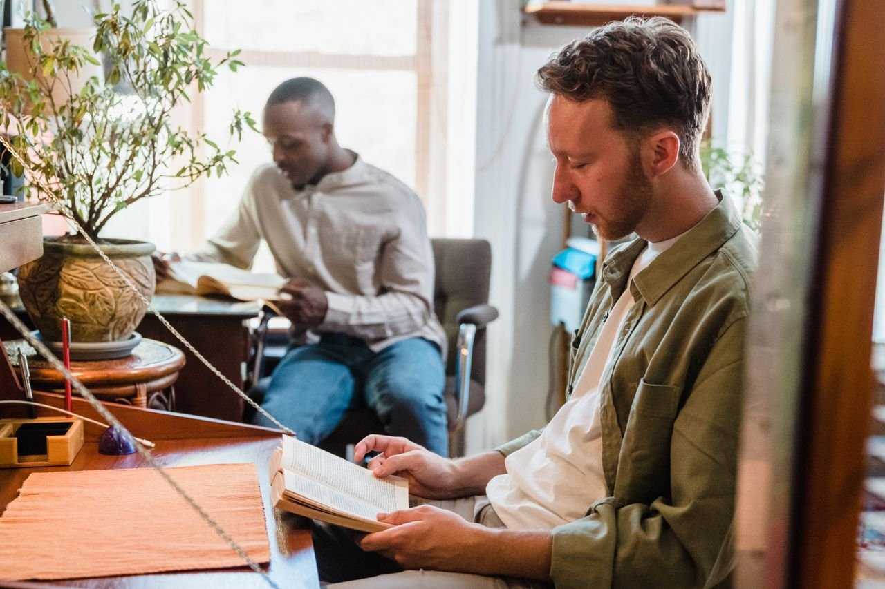 Two men enjoying a relaxing reading session in a bright, plant-filled living room.